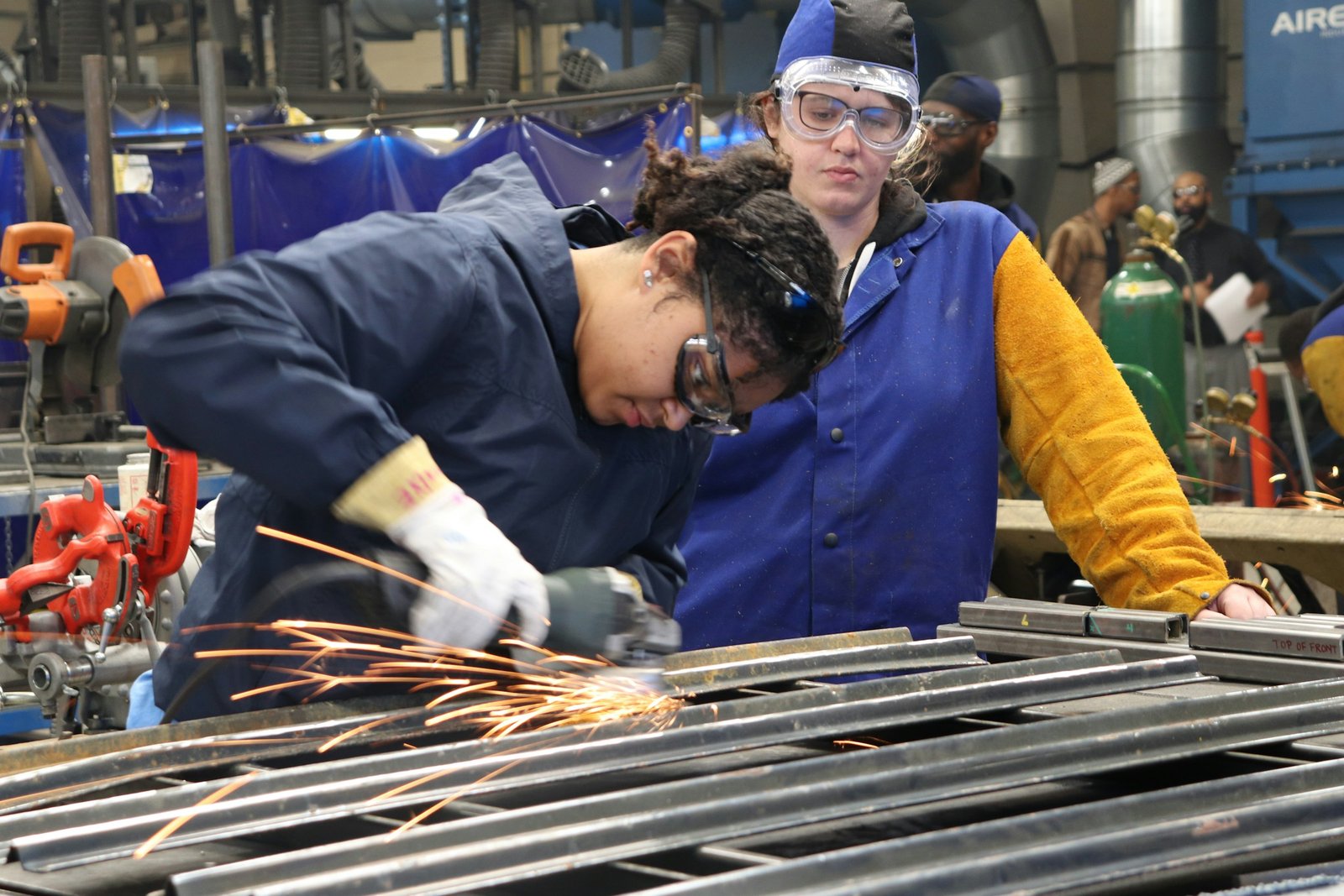 Two women in safety goggles doing metalwork at a workbench